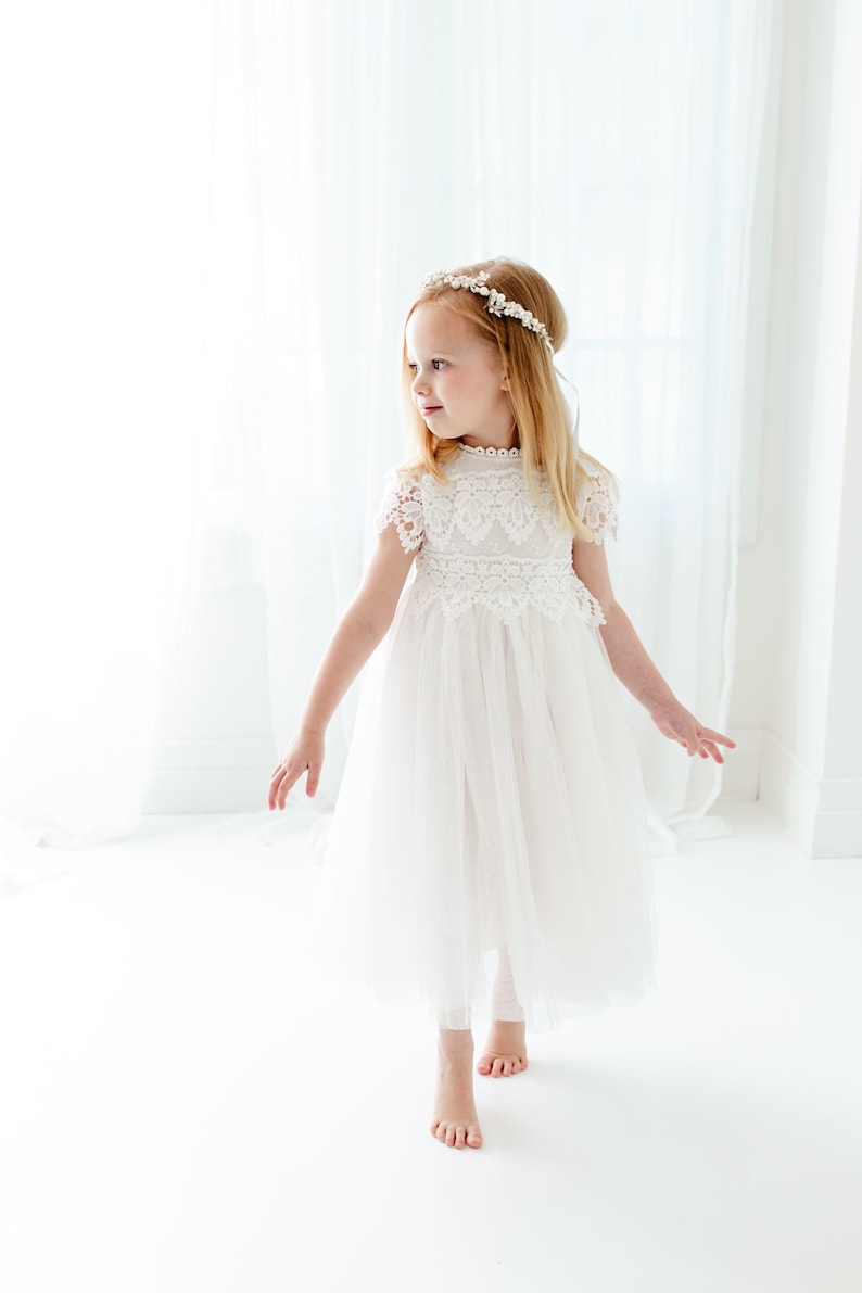 May include: A white dress featuring a lace bodice with short sleeves and a high neckline, paired with a layered tulle skirt. The garment is accessorized with a delicate floral headband featuring small white flowers and beads.