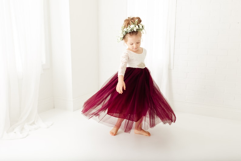 May include: A young girl wearing a white lace top with a burgundy tulle skirt and a flower crown. She is twirling in a white room.