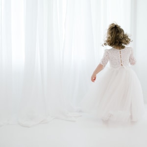May include: A young girl in a white lace dress with a tulle skirt stands in front of a white curtain. She is facing away from the camera and her hair is pulled back.