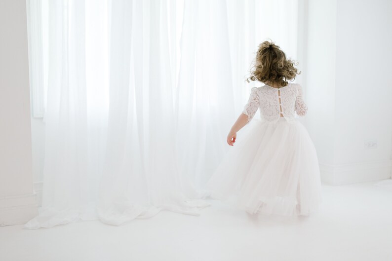May include: A young girl in a white lace dress with a tulle skirt stands in front of a white curtain. She is facing away from the camera and her hair is pulled back.