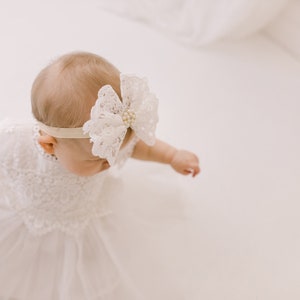 K&ouml;nnte beinhalten: Ein kleines M&auml;dchen in einem wei&szlig;en Spitzenkleid und einem wei&szlig;en Spitzenhaarband mit Perlenverzierung. Das Baby steht auf einer wei&szlig;en Oberfl&auml;che.