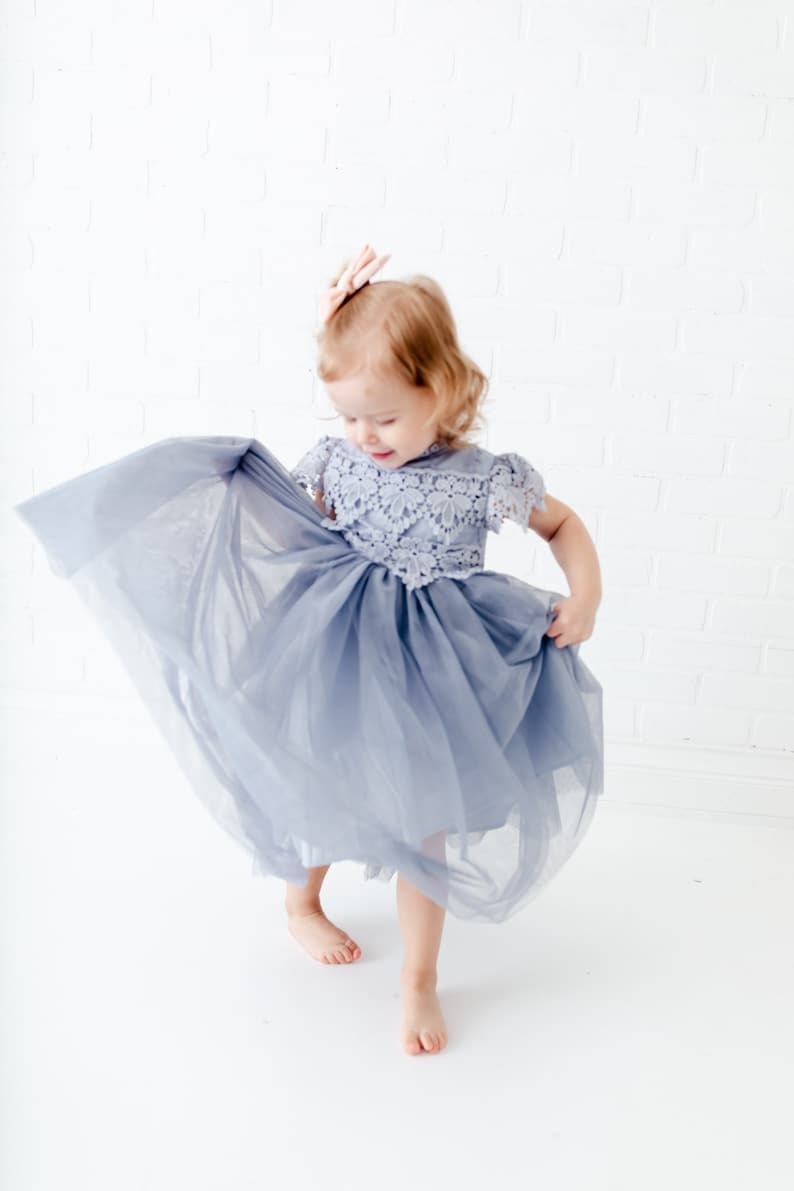 K&ouml;nnte beinhalten: Ein junges M&auml;dchen tr&auml;gt ein hellblaues T&uuml;llkleid mit Spitzendetails. Das Kleid hat einen vollen Rock und kurze &Auml;rmel. Das M&auml;dchen steht auf einem wei&szlig;en Hintergrund und h&auml;lt den Rock ihres Kleides.