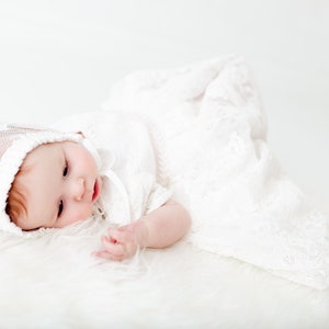 May include: A baby wearing a white lace bonnet and a matching white lace gown. The bonnet features intricate floral embroidery and a scalloped edge, while the long gown is crafted from delicate white lace fabric with a detailed floral pattern.