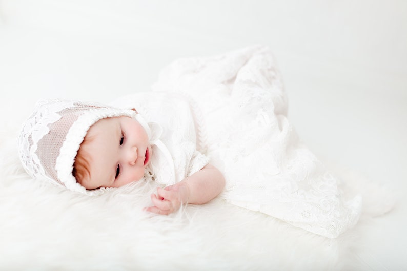 May include: A baby wearing a white lace bonnet and a matching white lace gown. The bonnet features intricate floral embroidery and a scalloped edge, while the long gown is crafted from delicate white lace fabric with a detailed floral pattern.