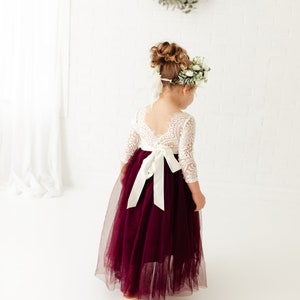 May include: A young girl wearing a white lace dress with long sleeves and a burgundy tulle skirt. The dress has a white satin sash tied in a bow at the back. The girl is wearing a flower crown and has her hair pulled back in a bun.