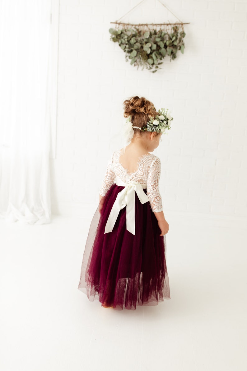 May include: A young girl wearing a white lace dress with long sleeves and a burgundy tulle skirt. The dress has a white satin sash tied in a bow at the back. The girl is wearing a flower crown and has her hair pulled back in a bun.