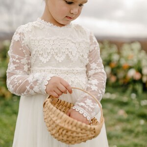 May include: A young girl in a white lace dress with long sleeves holds a woven basket. She is wearing a flower crown and looking down at the basket.
