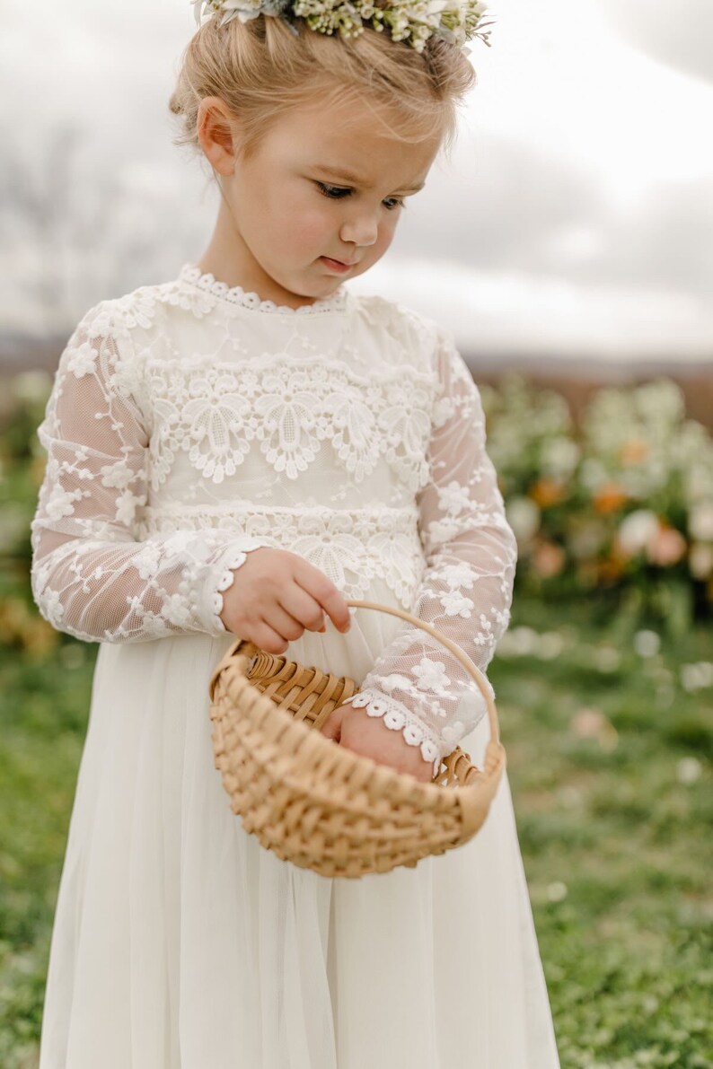 May include: A young girl in a white lace dress with long sleeves holds a woven basket. She is wearing a flower crown and looking down at the basket.