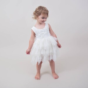 Op de afbeelding: Een jong meisje draagt een witte tule jurk met een kantboord. De jurk heeft meerdere lagen tule en een gerimpelde zoom.