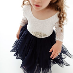 May include: A young girl wearing a white lace long-sleeved dress with a navy blue tulle skirt and a jeweled belt.