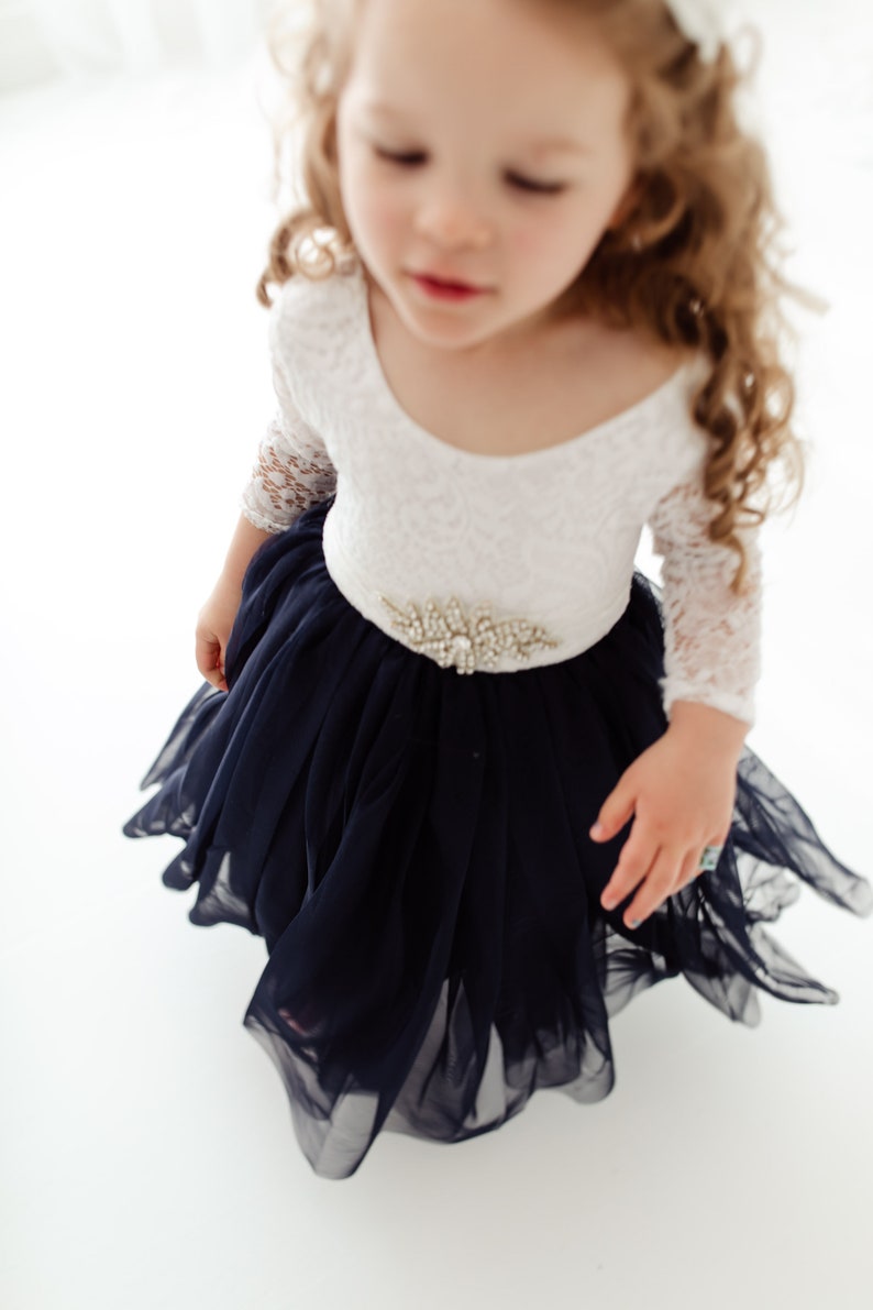 May include: A young girl wearing a white lace long-sleeved dress with a navy blue tulle skirt and a jeweled belt.