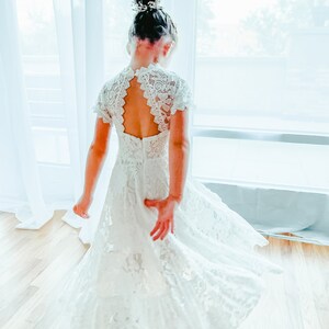 K&ouml;nnte beinhalten: Ein wei&szlig;es Spitzenkleid mit kurzen &Auml;rmeln, einem hochgeschlossenen Ausschnitt und einer tropfenf&ouml;rmigen R&uuml;cken&ouml;ffnung. Das Kleid hat ein k&ouml;rpernah geschnittenes Oberteil, das in einen weit schwingenden Rock &uuml;bergeht, verziert mit floralen Mustern.