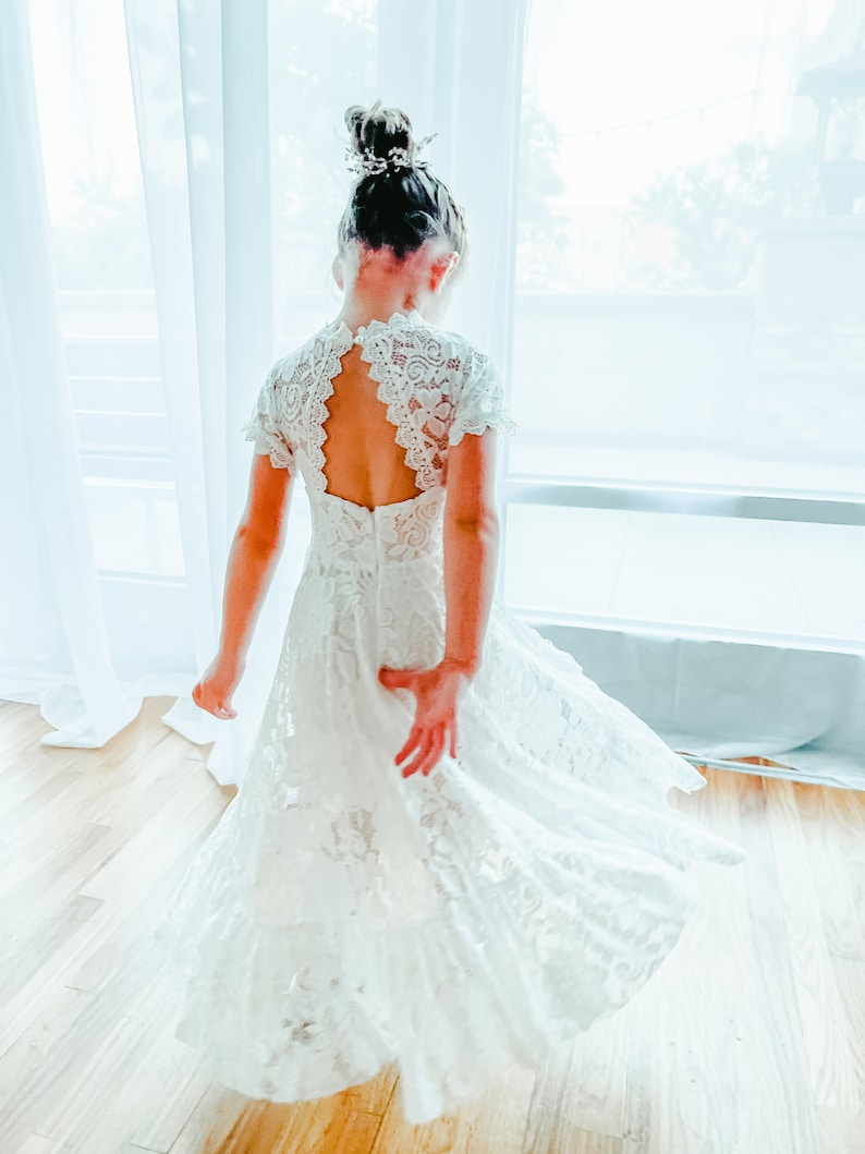 K&ouml;nnte beinhalten: Ein wei&szlig;es Spitzenkleid mit kurzen &Auml;rmeln, einem hochgeschlossenen Ausschnitt und einer tropfenf&ouml;rmigen R&uuml;cken&ouml;ffnung. Das Kleid hat ein k&ouml;rpernah geschnittenes Oberteil, das in einen weit schwingenden Rock &uuml;bergeht, verziert mit floralen Mustern.