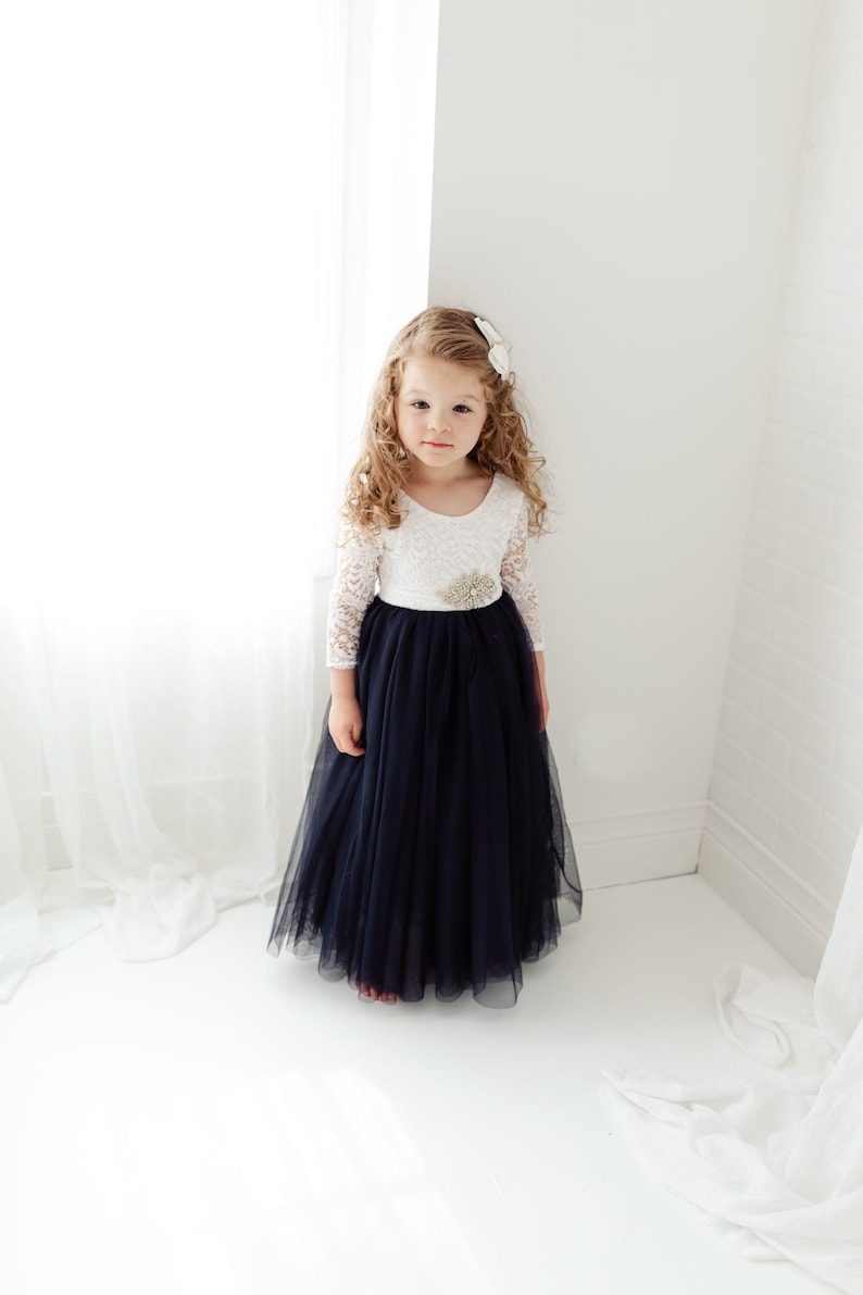 May include: A young girl wearing a white lace top with a navy blue tulle skirt and a sparkly flower embellishment on the waist.