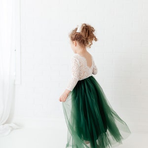 Pu&ograve; includere: Un abito lungo fino ai piedi con corpetto in pizzo bianco a maniche lunghe e scollo profondo sulla schiena, abbinato a una voluminosa gonna in tulle a strati color verde bosco scuro. Il capo presenta un punto vita aderente e una linea fluida.