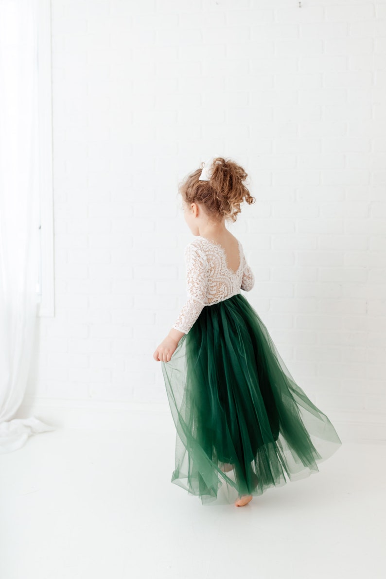 Pu&ograve; includere: Un abito lungo fino ai piedi con corpetto in pizzo bianco a maniche lunghe e scollo profondo sulla schiena, abbinato a una voluminosa gonna in tulle a strati color verde bosco scuro. Il capo presenta un punto vita aderente e una linea fluida.