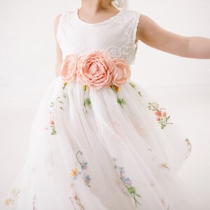Puede incluir: Un vestido de tul blanco con un corpi&ntilde;o de encaje y una faja de flores de melocot&oacute;n. El vestido est&aacute; bordado con peque&ntilde;as flores de varios colores.