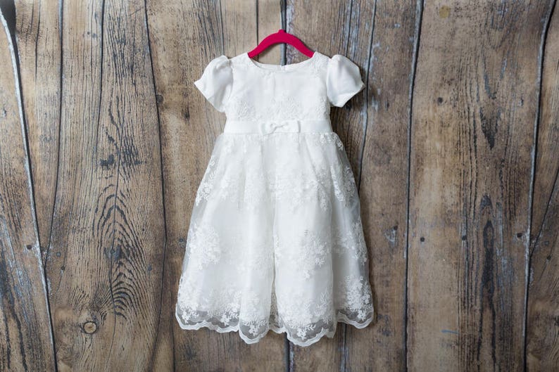 May include: A white dress featuring short puff sleeves, a round neckline, and a fitted bodice with a small bow at the waist. The skirt is made of a sheer overlay with intricate floral embroidery and a scalloped hem, hanging on a pink hanger.
