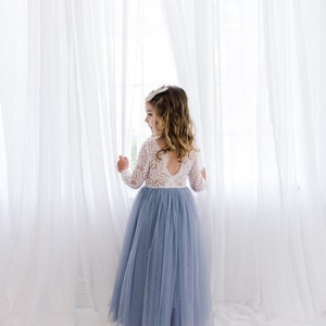May include: A young girl wearing a white lace long-sleeved top and a light blue tulle skirt stands in front of a white curtain.
