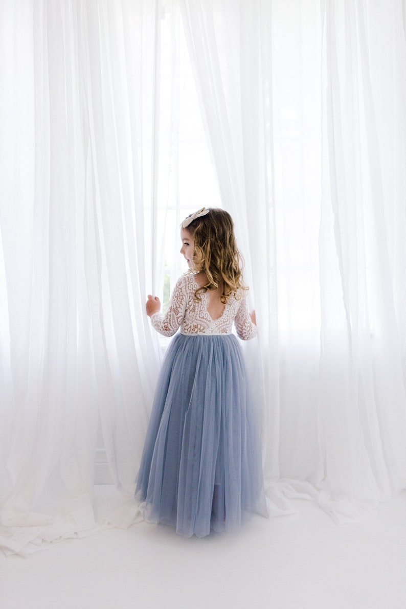 May include: A young girl wearing a white lace long-sleeved top and a light blue tulle skirt stands in front of a white curtain.