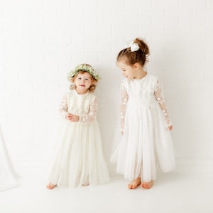 May include: Two young girls wearing white lace dresses. One girl is wearing a flower crown and the other has a white bow in her hair. They are standing in front of a white brick wall.