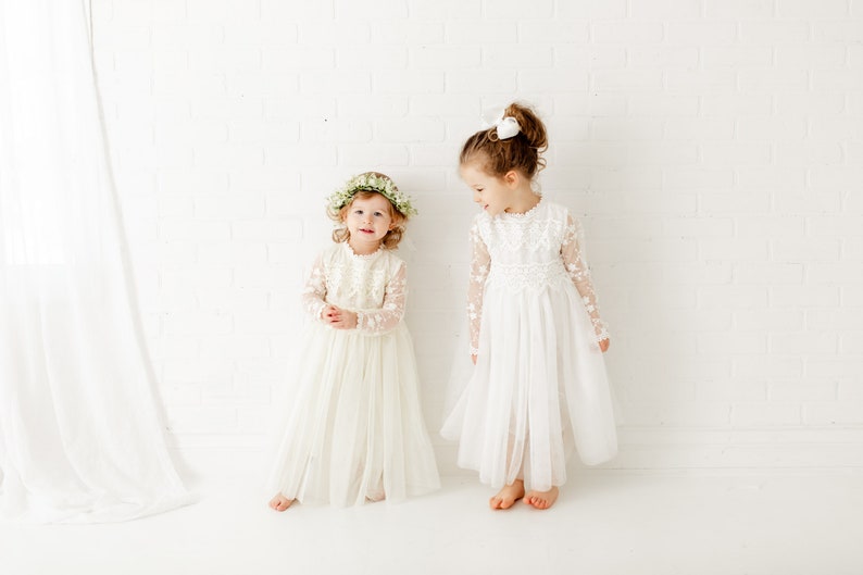 May include: Two young girls wearing white lace dresses. One girl is wearing a flower crown and the other has a white bow in her hair. They are standing in front of a white brick wall.