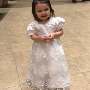 May include: A white floor-length dress with short puff sleeves and a round neckline. The bodice and skirt feature intricate white floral embroidery and lace overlays. A matching white bow hair accessory is worn in the dark hair of the child.