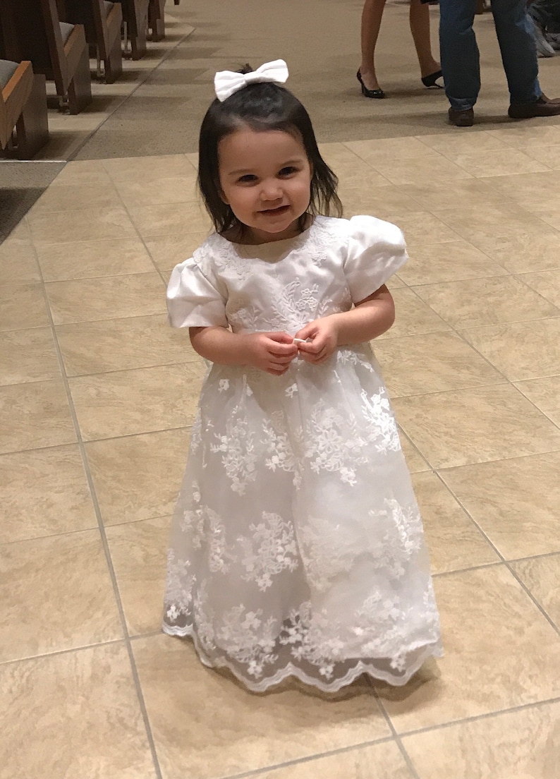 May include: A white floor-length dress with short puff sleeves and a round neckline. The bodice and skirt feature intricate white floral embroidery and lace overlays. A matching white bow hair accessory is worn in the dark hair of the child.