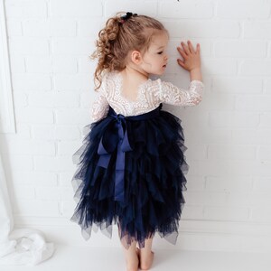May include: A dress featuring a white lace bodice with three-quarter length sleeves and a deep scoop back, paired with a voluminous navy blue tulle skirt. The waist is accented by a navy blue satin ribbon tied into a bow at the centre back.