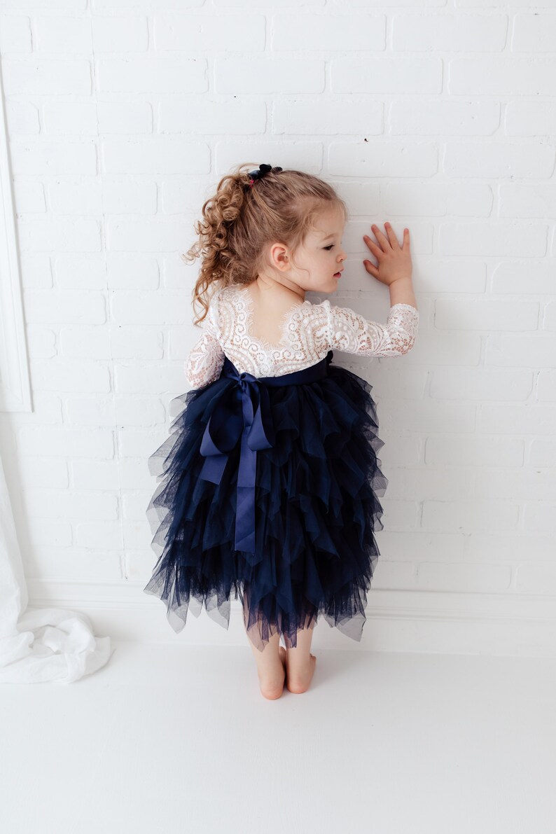 May include: A dress featuring a white lace bodice with three-quarter length sleeves and a deep scoop back, paired with a voluminous navy blue tulle skirt. The waist is accented by a navy blue satin ribbon tied into a bow at the centre back.