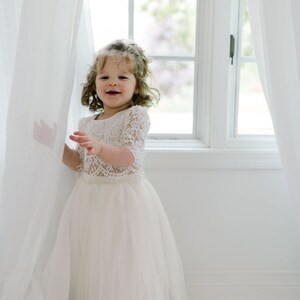 May include: A young child wearing a white lace top with short sleeves and a long, flowing tulle skirt. The child is smiling and standing near a window with white curtains. The dress appears to be a formal or special occasion outfit.