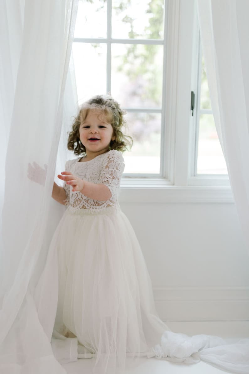 May include: A young child wearing a white lace top with short sleeves and a long, flowing tulle skirt. The child is smiling and standing near a window with white curtains. The dress appears to be a formal or special occasion outfit.
