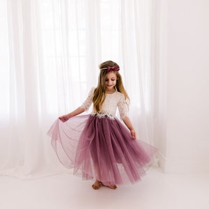 May include: A young girl wearing a white lace top and a long, flowing mauve tulle skirt. She is twirling in front of a white backdrop.