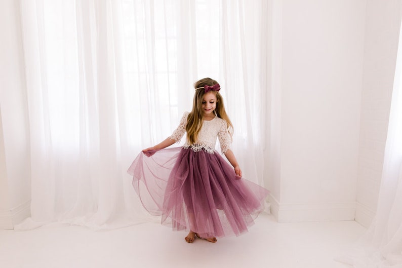 May include: A young girl wearing a white lace top and a long, flowing mauve tulle skirt. She is twirling in front of a white backdrop.