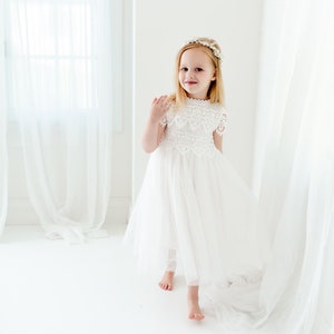 May include: A white dress featuring a lace bodice with short sleeves and a high neckline, paired with a layered tulle skirt. The garment is accessorized with a floral headpiece featuring small white flowers and green leaves on a thin band.