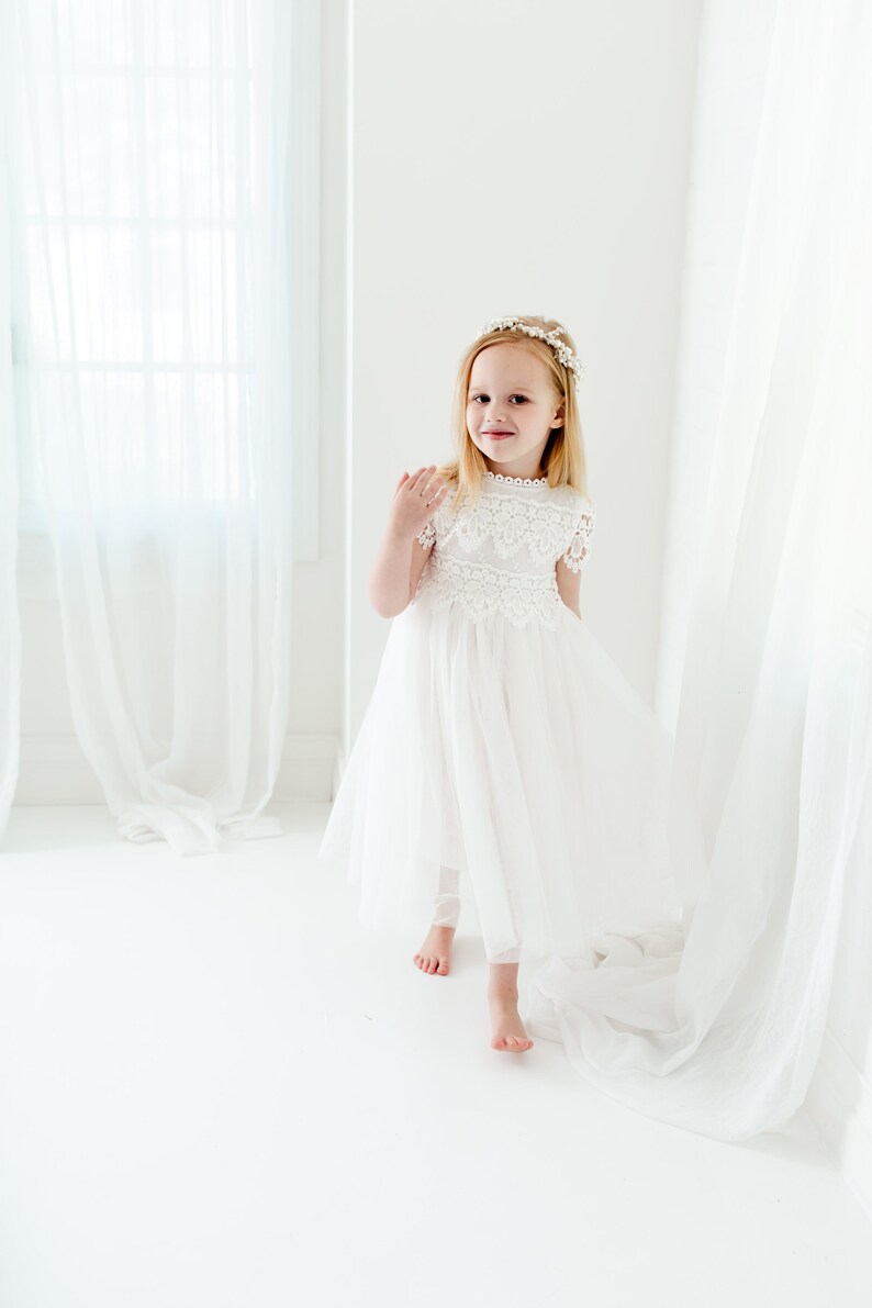 May include: A white dress featuring a lace bodice with short sleeves and a high neckline, paired with a layered tulle skirt. The garment is accessorized with a floral headpiece featuring small white flowers and green leaves on a thin band.