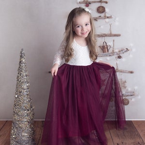 May include: A young girl wearing a white lace top and a burgundy tulle skirt. She is standing in front of a rustic Christmas tree made of branches and ornaments.