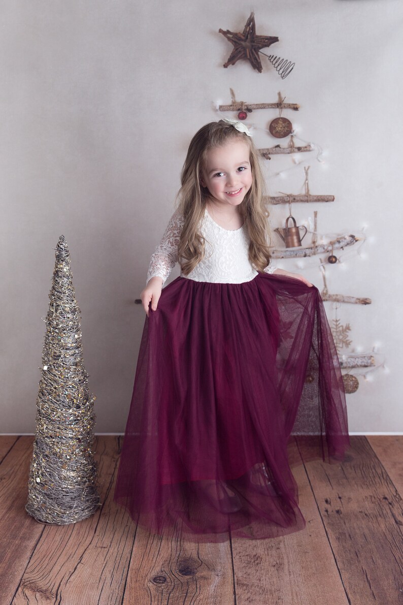 May include: A young girl wearing a white lace top and a burgundy tulle skirt. She is standing in front of a rustic Christmas tree made of branches and ornaments.