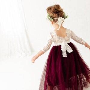 May include: A young girl wearing a white lace bodice dress with a burgundy tulle skirt. The dress has a lace-up back and a white bow. The girl is wearing a flower crown.
