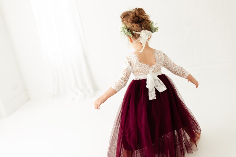 May include: A young girl wearing a white lace bodice dress with a burgundy tulle skirt. The dress has a lace-up back and a white bow. The girl is wearing a flower crown.