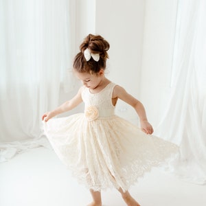 May include: A young girl wearing a white lace dress with a peach-colored flower accent at the waist. She is twirling in the dress and has a white bow in her hair.