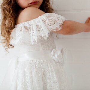 May include: A white off-the-shoulder dress with lace detailing and a satin ribbon sash. The dress has a full skirt and a flower accent on the side.