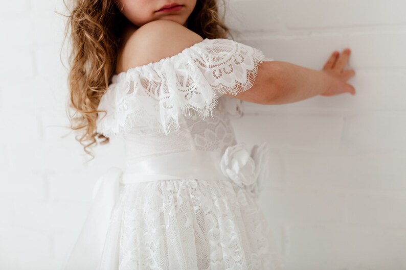 May include: A white off-the-shoulder dress with lace detailing and a satin ribbon sash. The dress has a full skirt and a flower accent on the side.