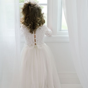 May include: A young person wearing a white lace dress with long sleeves and a tulle skirt, standing near a window. The dress features a button-up back. The person is looking out the window, with their back to the camera.