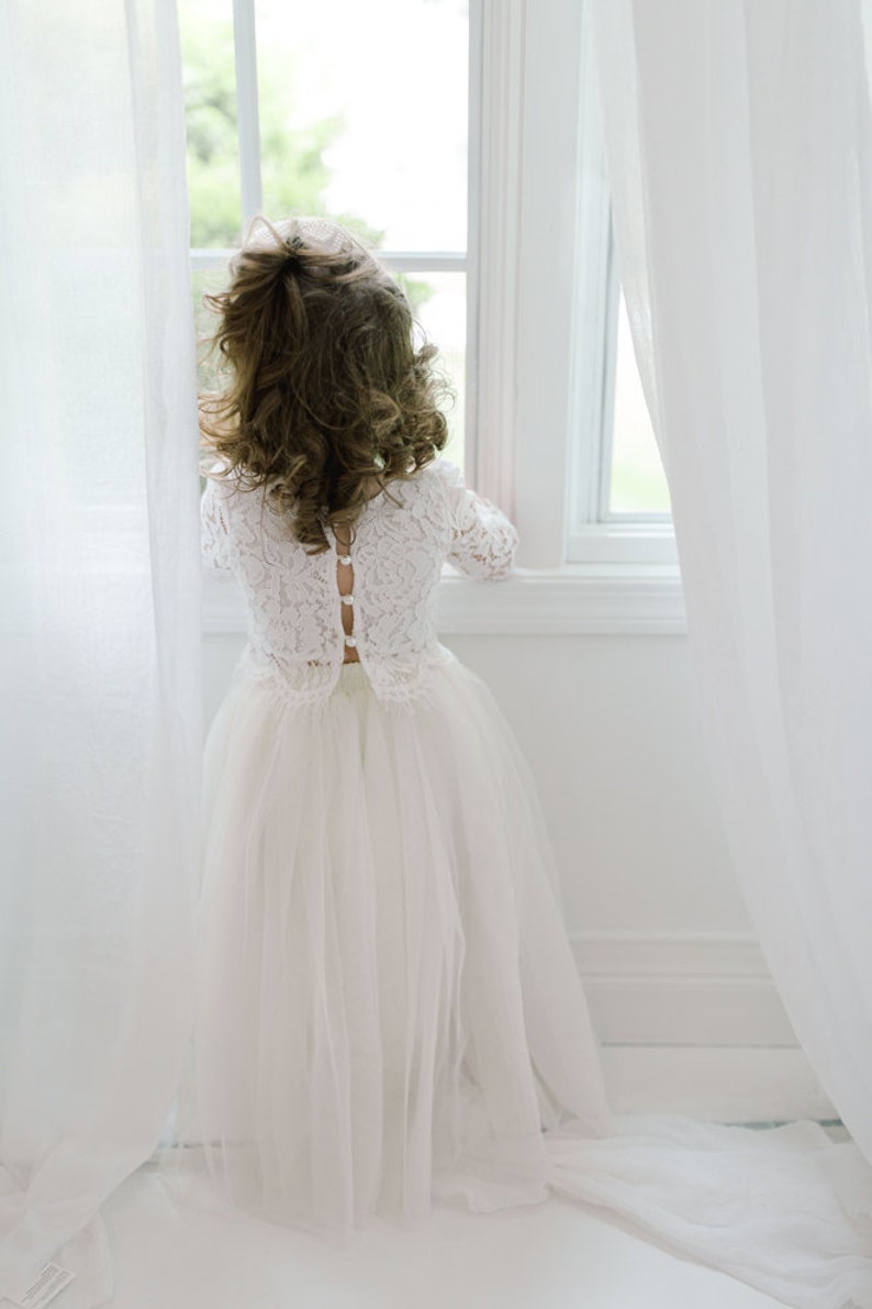 May include: A young person wearing a white lace dress with long sleeves and a tulle skirt, standing near a window. The dress features a button-up back. The person is looking out the window, with their back to the camera.