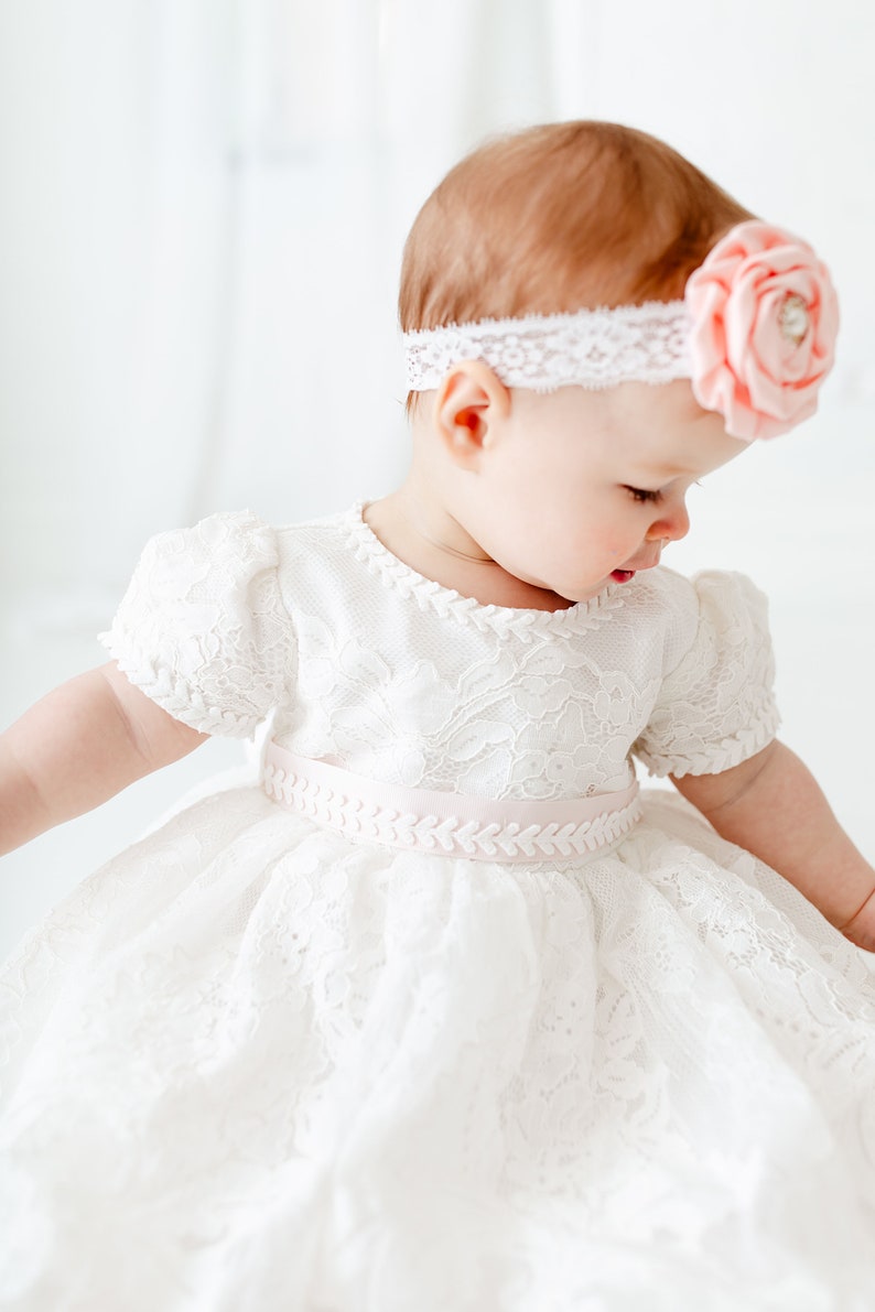 Puede incluir: Una ni&ntilde;a peque&ntilde;a con un vestido blanco de encaje con una cinta de raso rosa y una diadema de encaje blanco con una flor rosa.