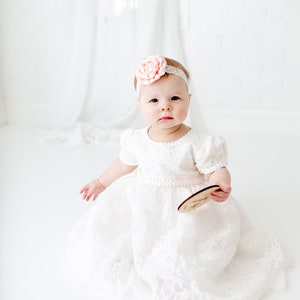 May include: A white lace dress with short puff sleeves and a pale pink ribbon waistband. The garment is paired with a matching white lace headband featuring a large, pale pink fabric flower centered on the front. The dress has a full skirt.
