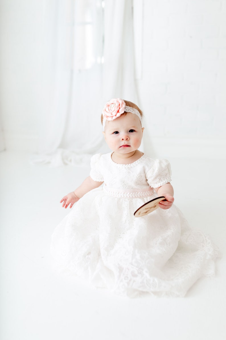 May include: A white lace dress with short puff sleeves and a pale pink ribbon waistband. The garment is paired with a matching white lace headband featuring a large, pale pink fabric flower centered on the front. The dress has a full skirt.