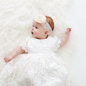 K&ouml;nnte beinhalten: Ein Baby M&auml;dchen in einem wei&szlig;en Spitzenkleid und einem Haarreif mit einer Pfirsichbl&uuml;te. Das Baby liegt auf einem wei&szlig;en, flauschigen Teppich.