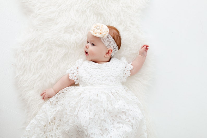 K&ouml;nnte beinhalten: Ein Baby M&auml;dchen in einem wei&szlig;en Spitzenkleid und einem Haarreif mit einer Pfirsichbl&uuml;te. Das Baby liegt auf einem wei&szlig;en, flauschigen Teppich.